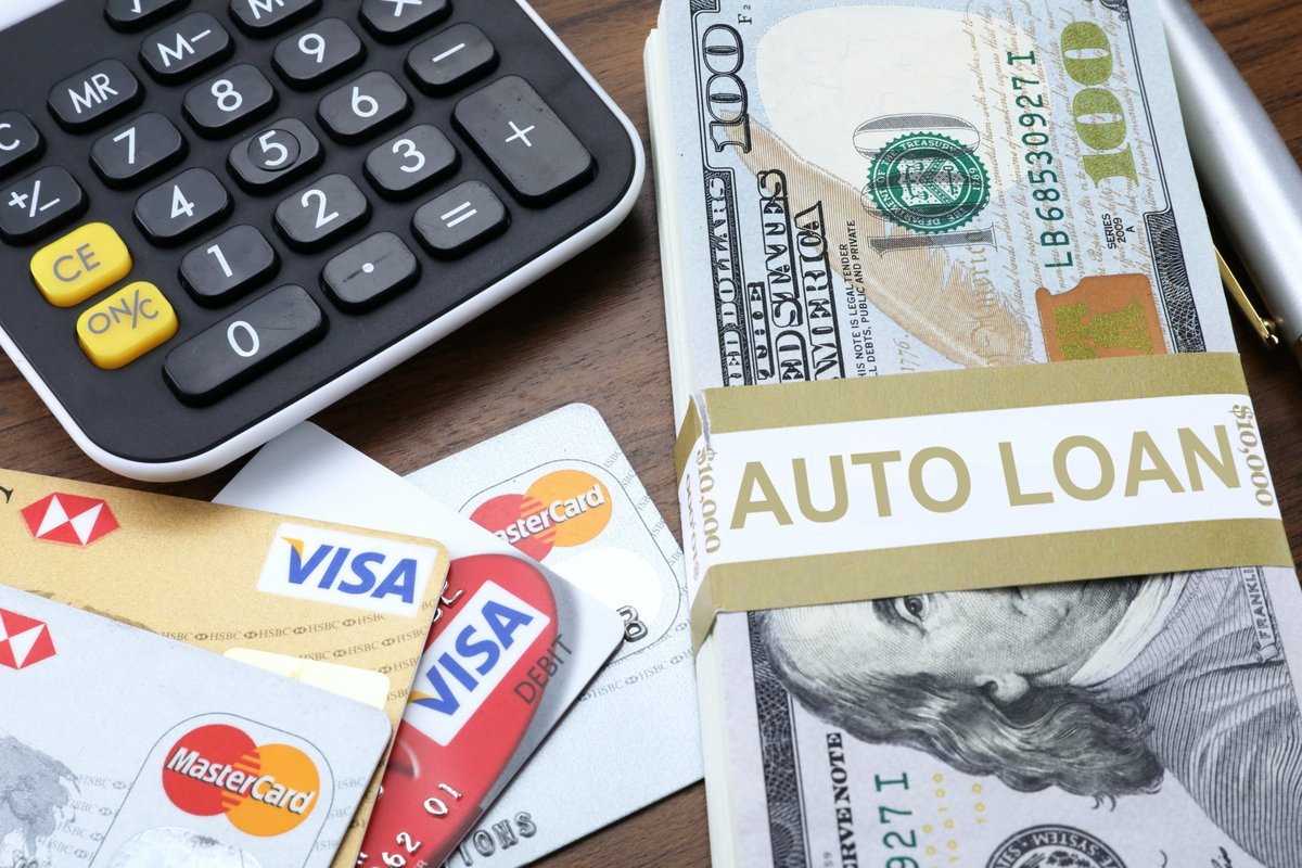 A calculator next to a stack of $100 bills wrapped in a gold band labeled AUTO LOAN, with Visa and Mastercard credit cards fanned out beside it on a dark wood desk